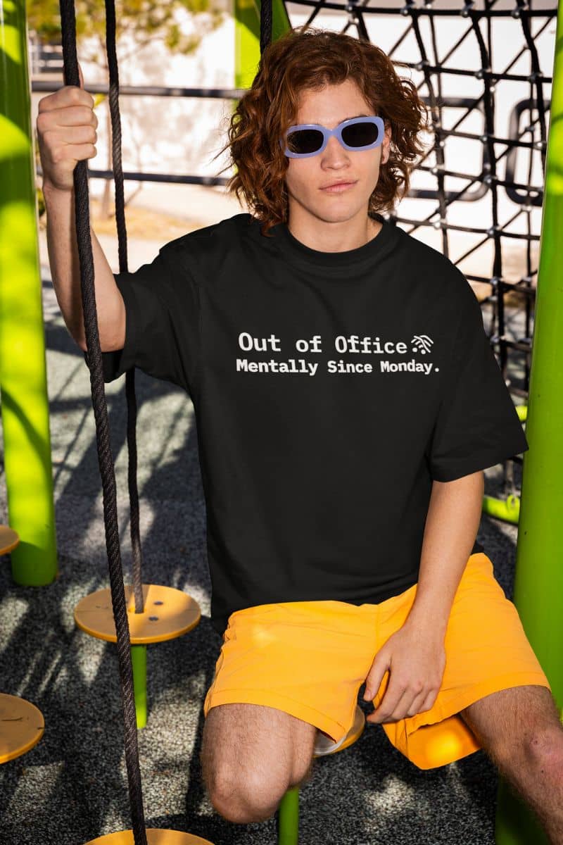 relatable job quote shirt with a person wearing a black t-shirt with text 'Out of Office. Mentally Since Monday.' and yellow shorts sitting on a swing.