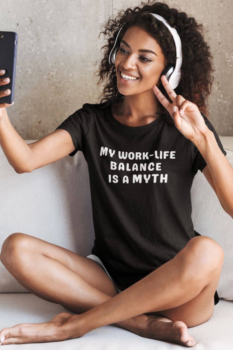 relatable work shirt with a woman wearing a black t-shirt with 'My work-life balance is a myth' text, sitting on a couch.
