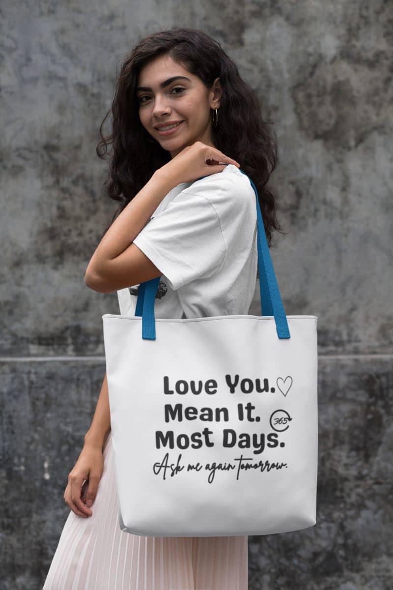 relationship quote tote with a woman holding a tote bag with text 'Love You. Mean It. Most Days. Ask me again tomorrow.' against a textured wall.