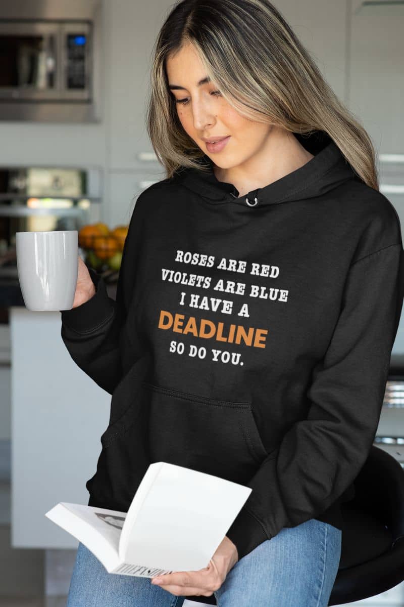 remote work hoodie with a woman wearing a black hoodie with text 'Roes are red, violets are blue. I have a deadline so you do', holding a mug and reading a book in a kitchen.