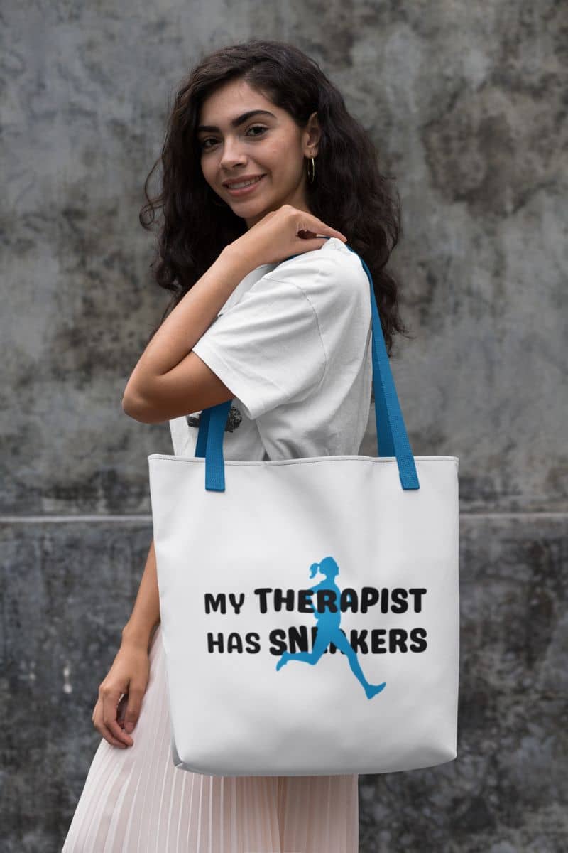 runner tote bag with a woman holding a tote bag with text 'My Therapist Has Sneakers' against a gray wall.