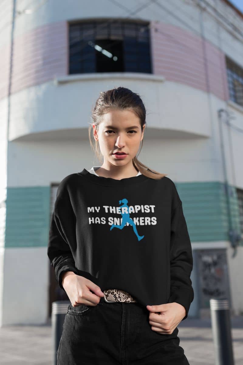 running quote sweatshirt with a woman wearing a black sweatshirt with text 'My Therapist Has Sneakers' and graphic, standing in front of a building.