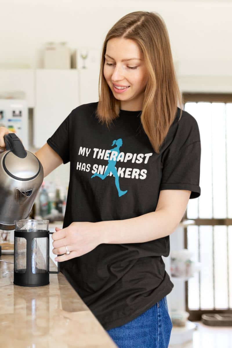 running quote t-shirt with a woman in a kitchen wearing a black t-shirt with text 'My Therapist Has Sneakers', holding a kettle.
