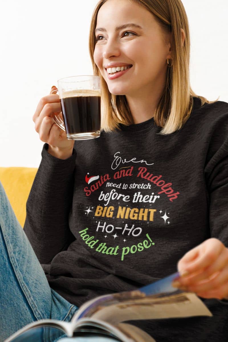 Santa and Rudolph sweatshirt featuring a woman wearing a sweatshirt with the text “Even Santa & Rudolph Need to Stretch Before Their Big Night. Ho-Ho-Hold That Pose!” while reading a magazine and holding a cup of coffee.