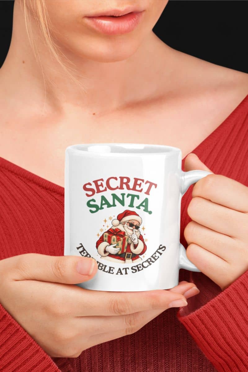 Santa Claus coffee mugs with a Secret Santa Terrible at Secrets design on a white ceramic mug held by a woman in a red sweater.