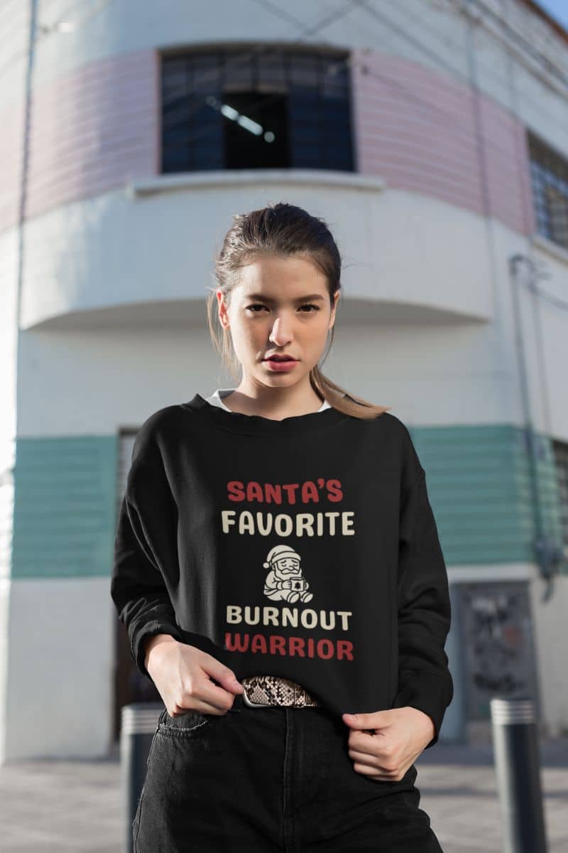 santa coffee lover sweater with a person wearing a black sweatshirt with text 'Santa’s Favorite Burnt Out Worrior' and graphics, standing in front of a building.