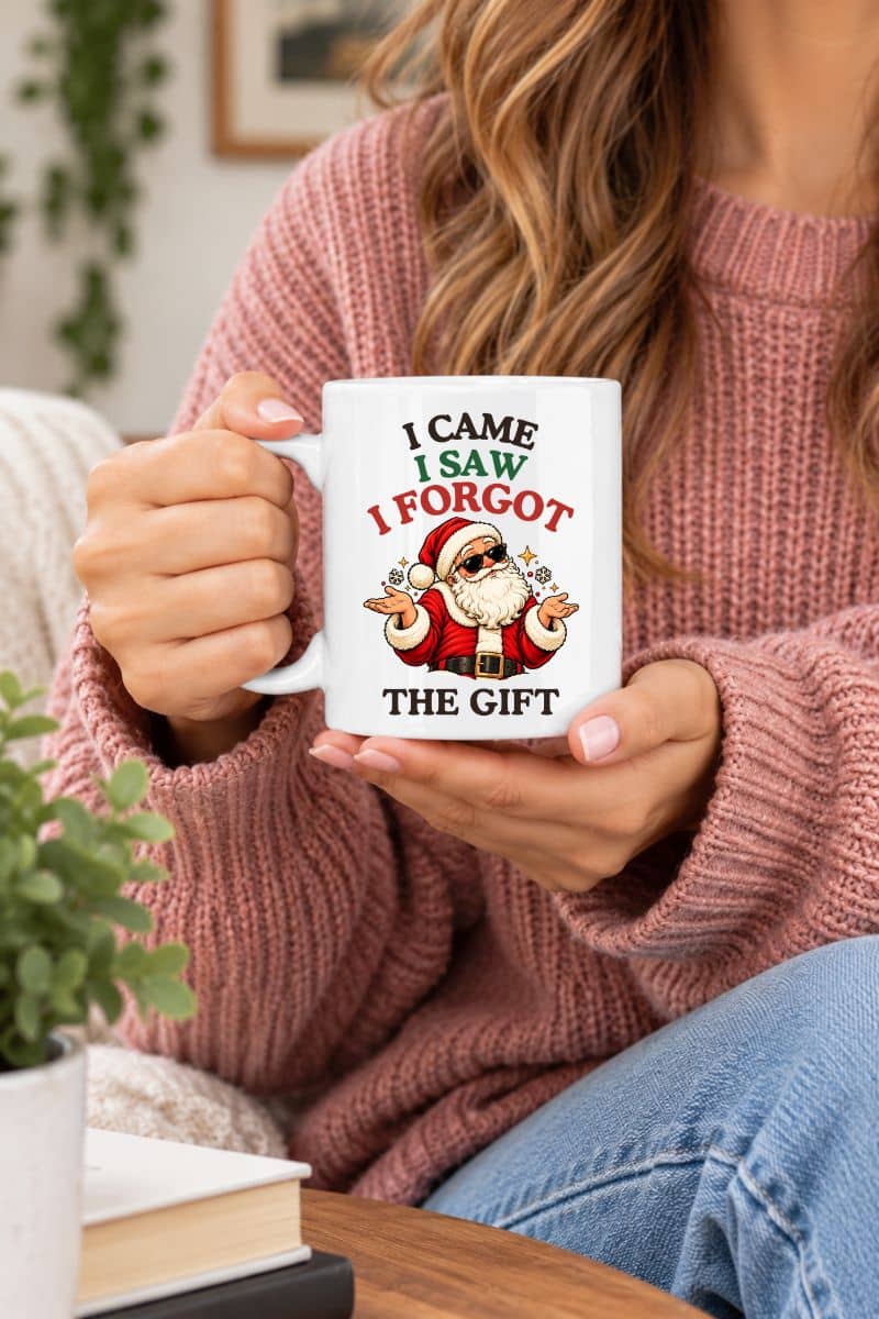 Woman in a pink knit sweater holding a Santa coffee mug featuring a shrugging Santa and “I Came. I Saw. I Forgot the Gift” design.