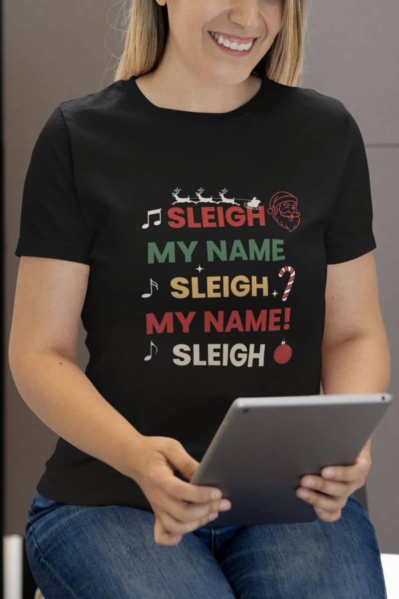 santa humor shirt with a person wearing a black t-shirt with colorful text 'Sleigh My Name, Sleigh My Name' and graphics, holding a tablet.