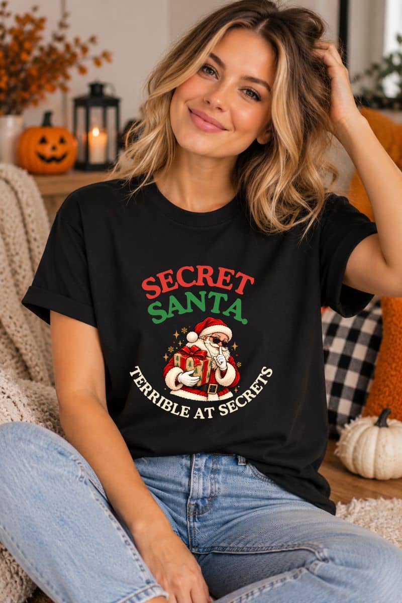 Santa shirt featuring the “Secret Santa. Terrible at Secrets.” design on a black tee worn by a smiling woman sitting indoors.