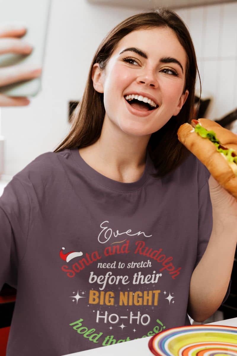 santa stretching shirt with a woman wearing a graphic t-shirt with text 'Event Santa & Rudolph Need to Stretch Before their Big Night. Ho-Ho-Hold That Pose!', holding a hot dog, indoors.