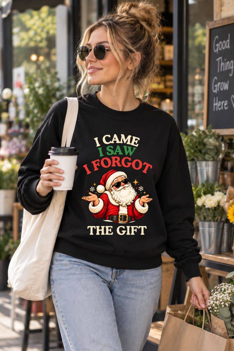 Woman carrying coffee and shopping bags in a black Santa sweatshirt featuring a shrugging Santa and the funny “I Came. I Saw. I Forgot the Gift” quote.
