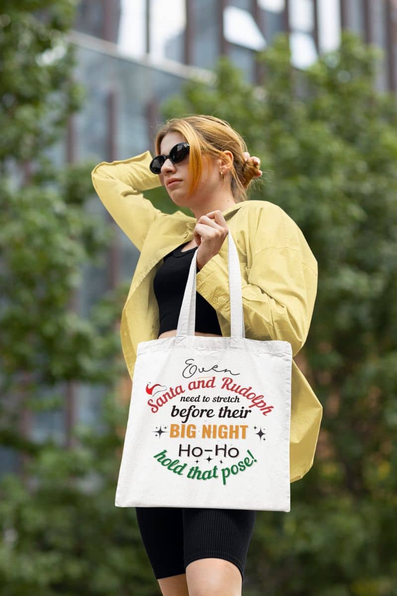 santa tote bag with the blonde girl posing with a tote bag that reads 'Event Santa & Rudolph Need to Stretch Before Their Big Night. Ho-Ho-Hold That Pose!'