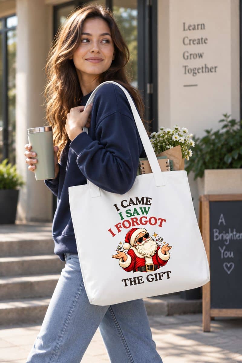 Woman in a navy sweatshirt carrying a Santa tote bag with a funny shrugging Santa design, flowers, and a notebook while holding a travel cup.
