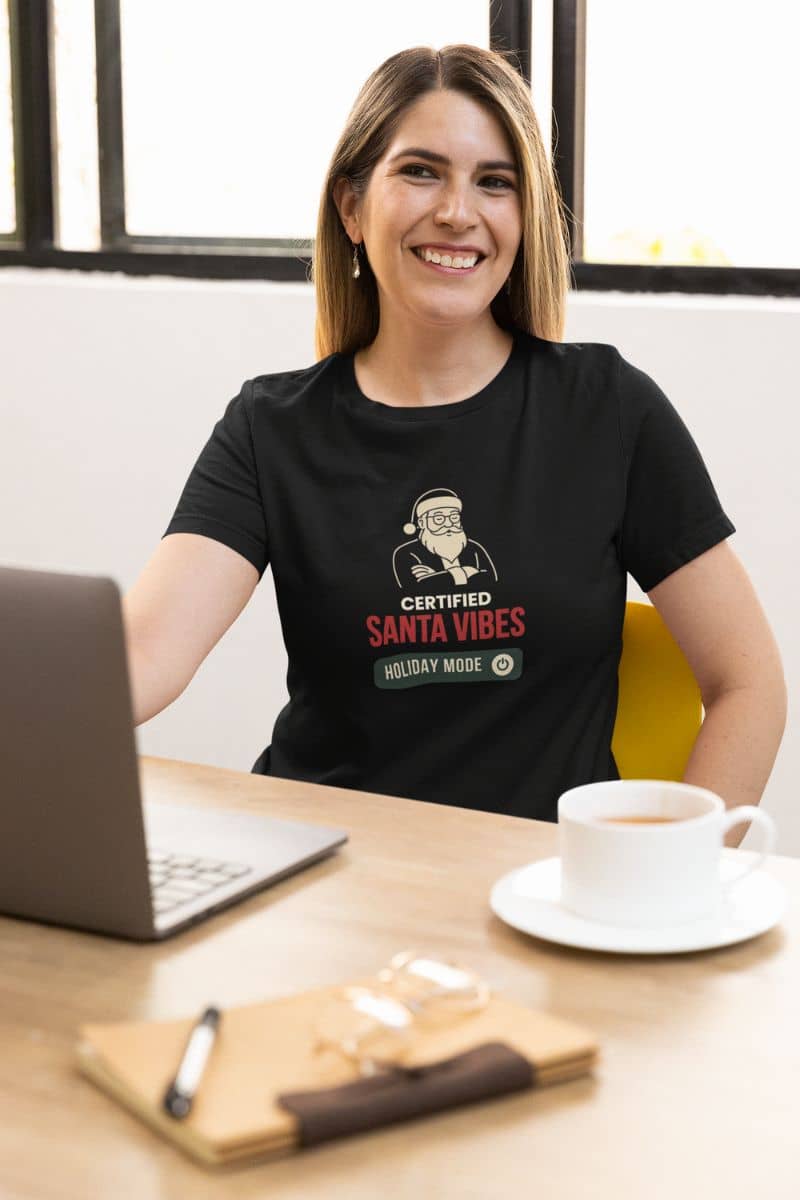 santa vibes t-shirt with a woman wearing a black t-shirt featuring the text 'Certified Santa Vibes – Holiday Mode On', sitting at a desk with a laptop and coffee.
