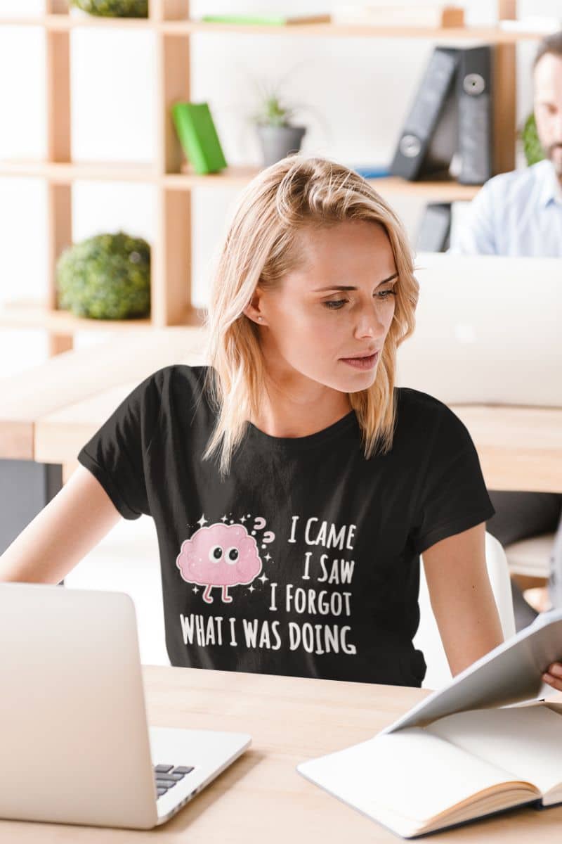 sarcastic graphic tee with a woman wearing a black t-shirt with text 'I came, I saw, I forgot what I was doing', sitting at a desk in an office setting.