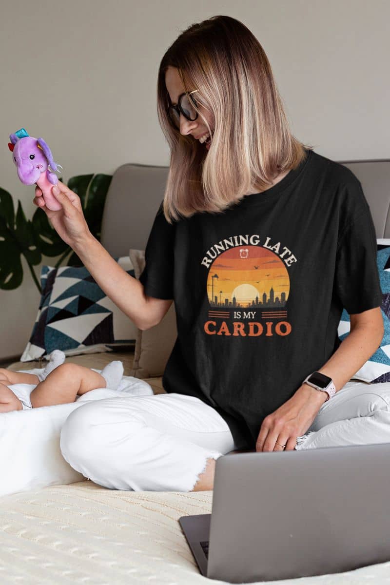 sarcastic graphic tee with a woman wearing a black t-shirt with text 'Running late is my cardio', sitting on a bed with a laptop and phone.