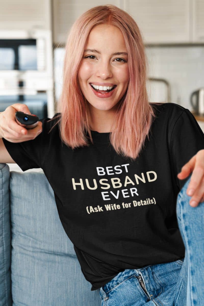 sarcastic husband shirt featuring a woman  wearing a t-shirt with text 'Best Husband Ever. (Ask Wife for Details)' while watching TV with a remote control