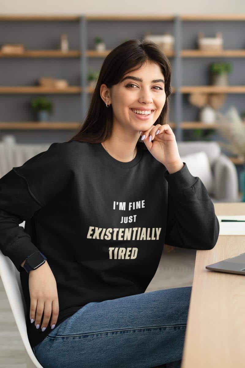 sarcastic introvert pullover with a woman wearing a black sweatshirt with text 'I’m fine. Just existentially tired.' , sitting in an office setting.