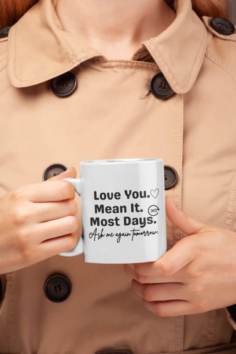 sarcastic love mug with a person holding a mug with text 'Love You. Mean It. Most Days. Ask me again tomorrow.' in a beige coat