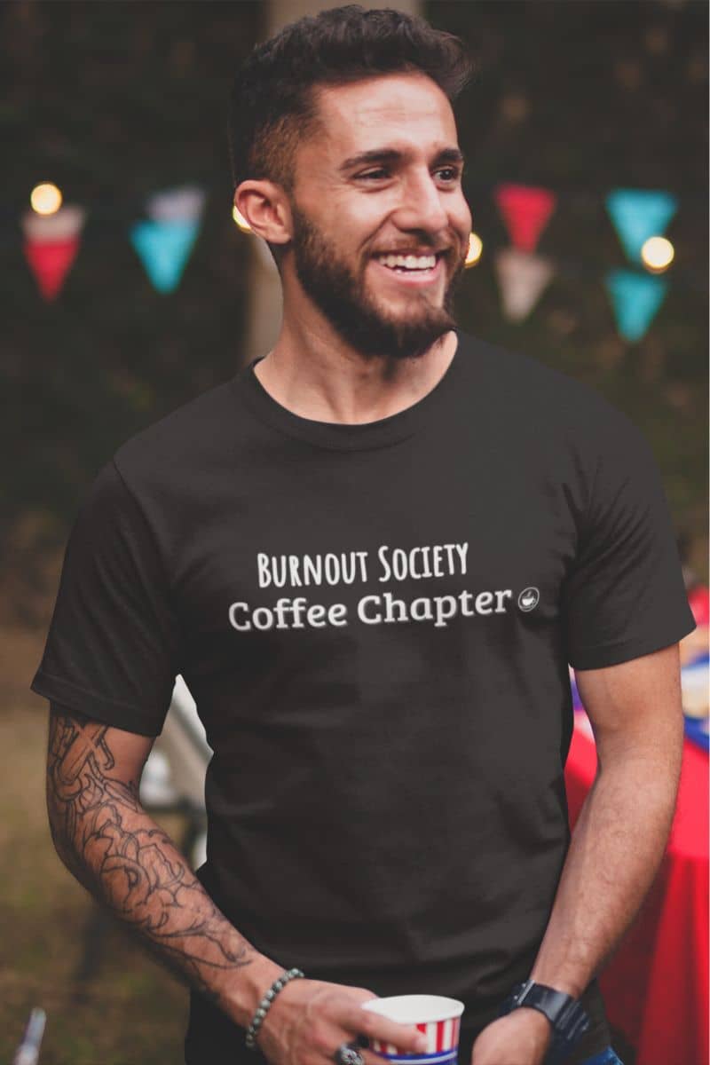 sarcastic mental health tshirt with a man wearing a black t-shirt with 'Burnout Society Coffee Chapter' text, standing outdoors with colorful flags in the background.