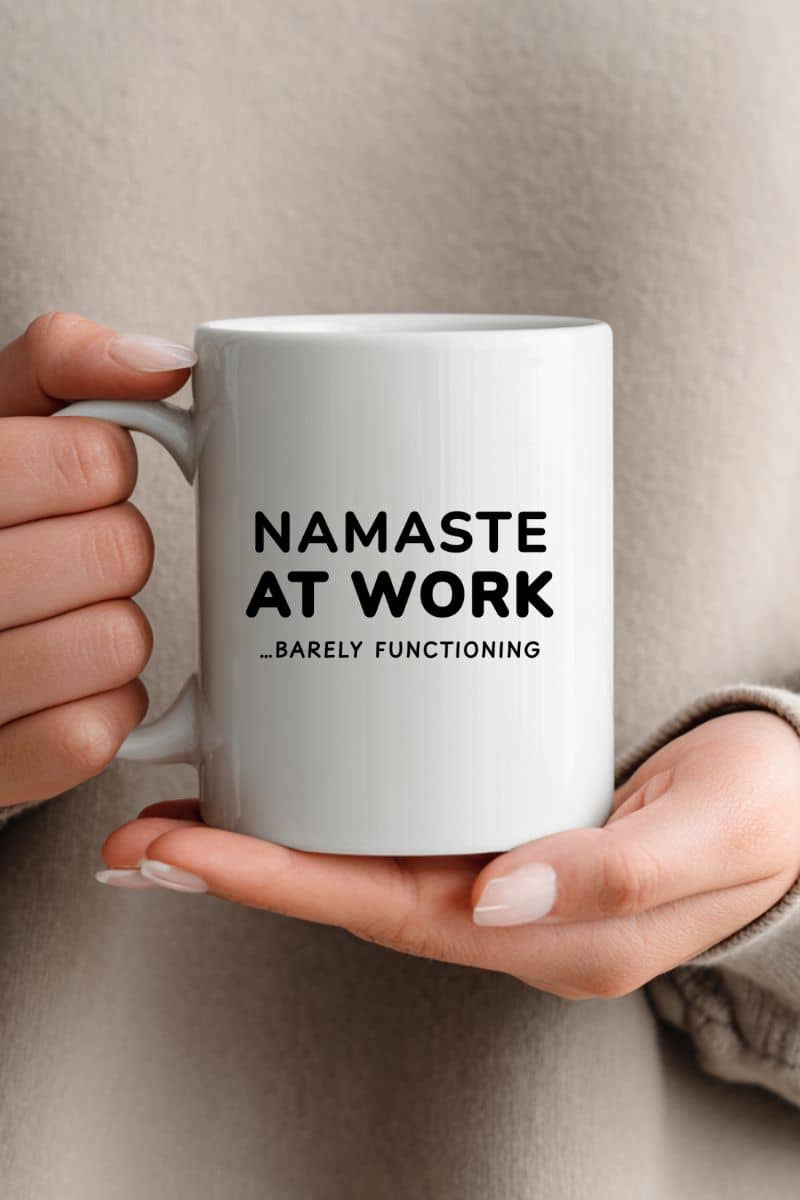 sarcastic mug with a person holding a white mug with text 'Namaste at Work...barely functioning' on a neutral background