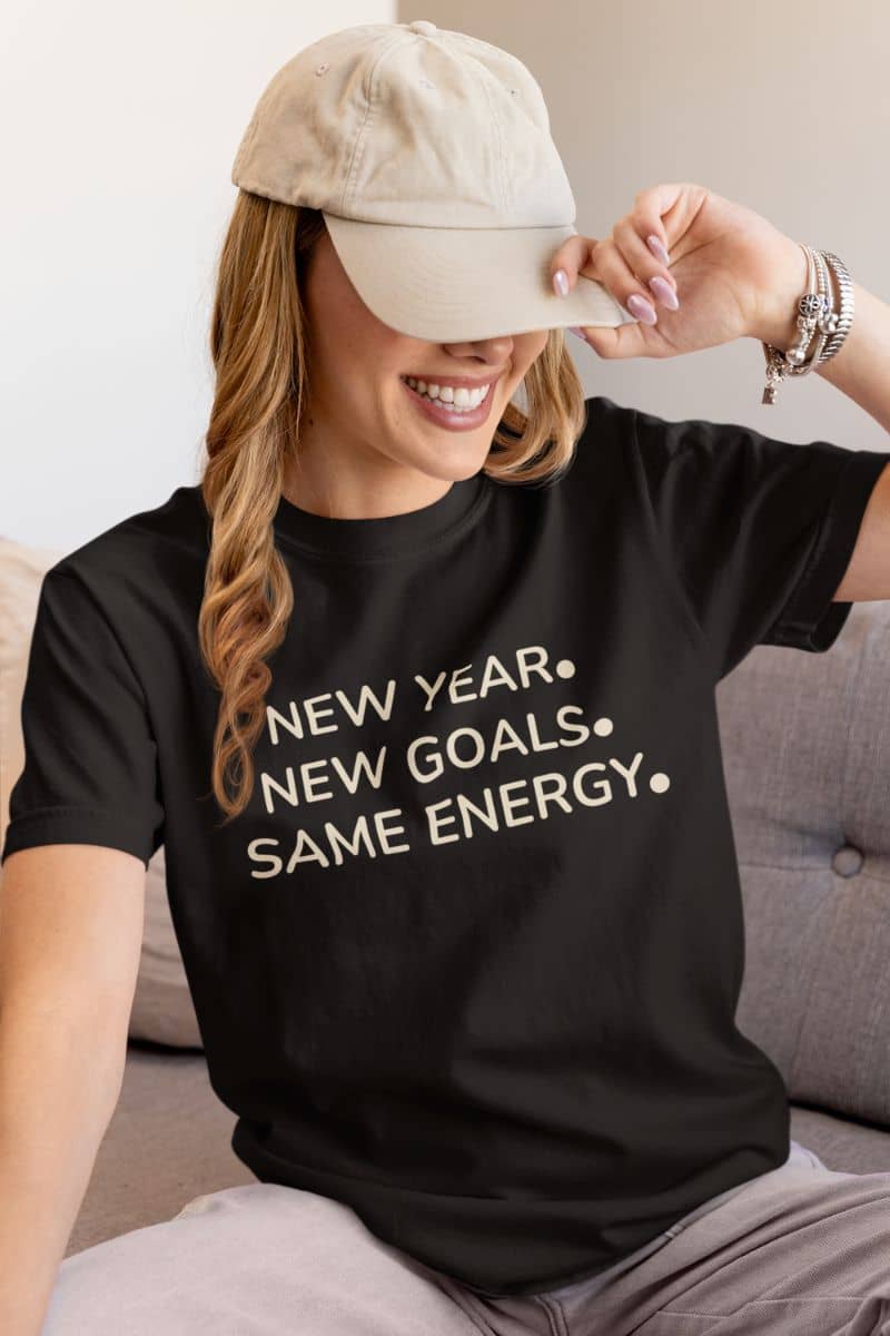 sarcastic new year shirt with a person wearing a black t-shirt with 'New Year. New Goals. Same Energy.' text, sitting on a couch.