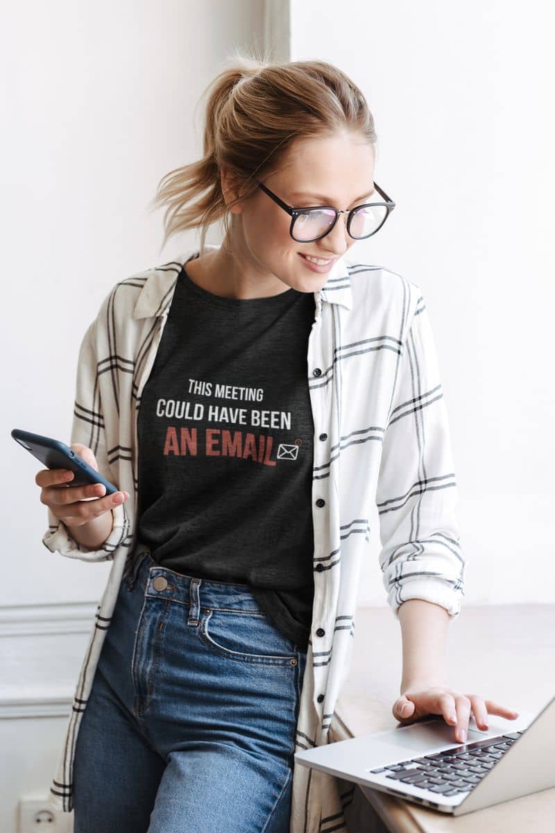 sarcastic office shirt with a woman wearing a black t-shirt with text 'This Meeting Could Have Been An Email', using a laptop and phone indoors.