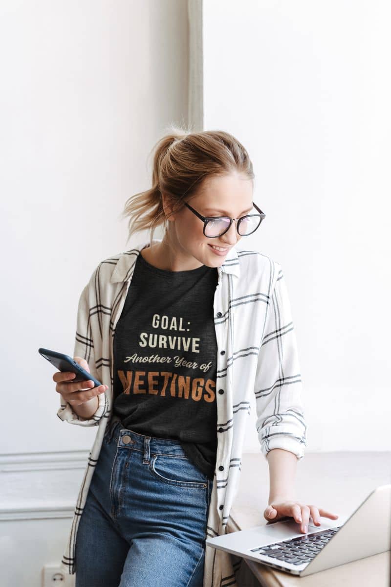 sarcastic office shirt with a woman wearing a black t-shirt with text 'Goal: Survive Another Year of Meetings', using a laptop.