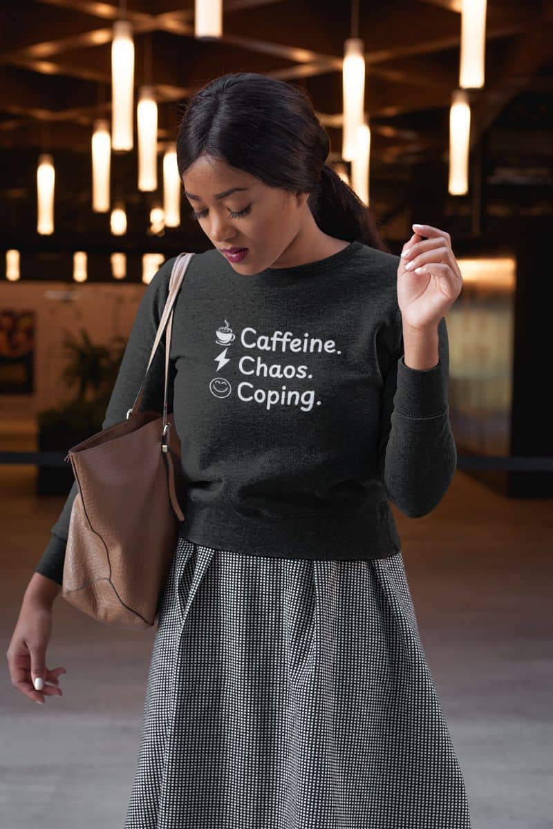 sarcastic self-care sweater with a woman wearing a black sweater with text 'Caffeine, Chaos & Coping', holding a brown bag in an indoor setting.