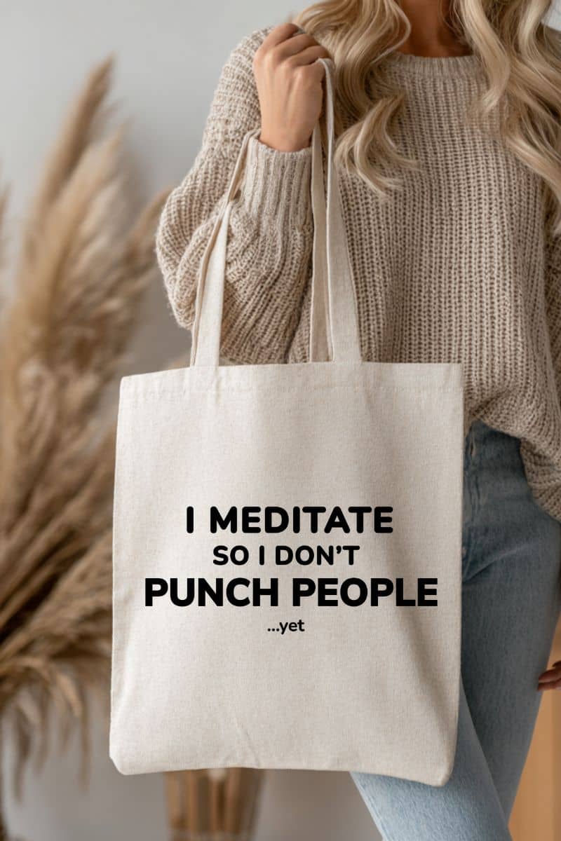 sarcastic tote bag with a person holding a tote bag with text 'I Meditate so I don't punch people... yet' against a neutral background.