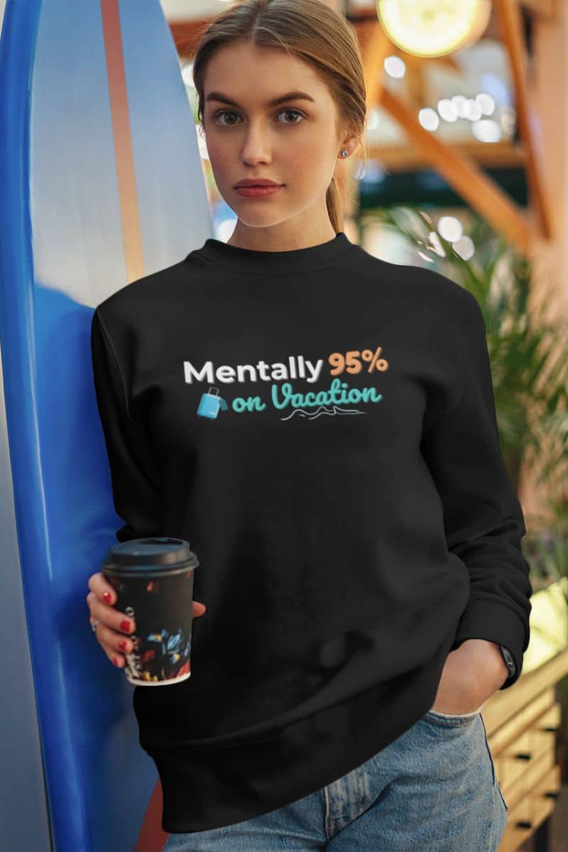 sarcastic vacation sweatshirt with a woman wearing a black sweatshirt with text 'Mentally 95% on Vacation', holding a coffee cup, standing next to a blue surfboard.