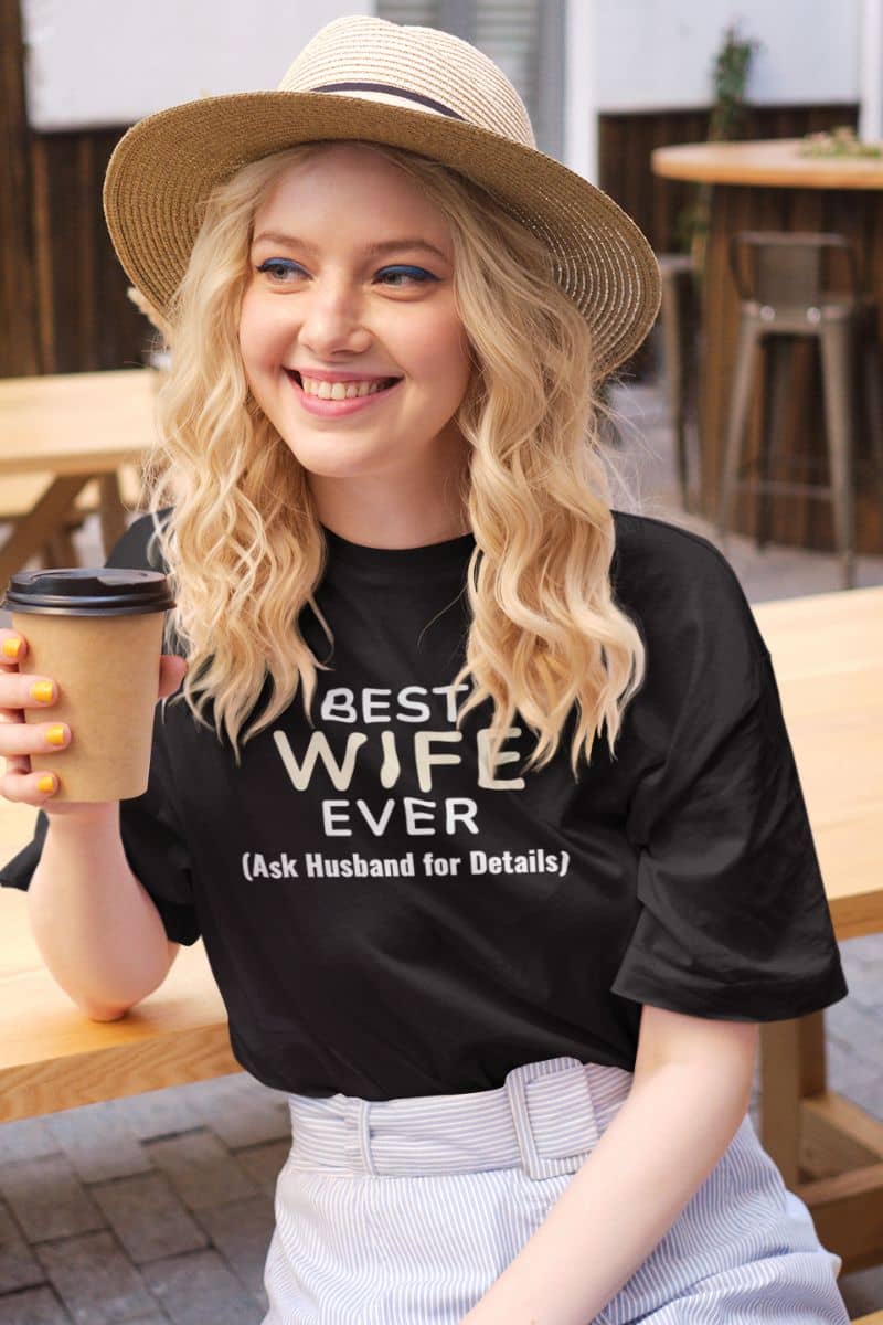 sarcastic wife tee with a woman wearing a black t-shirt with text 'Best Wife Ever. (Ask Husband for Details)', holding a coffee cup outdoors.