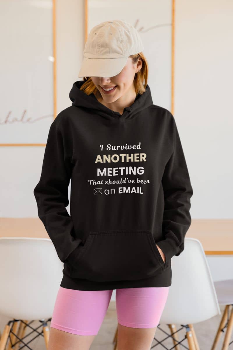 sarcastic work hoodie with a person wearing a black hoodie with text 'I Survived Another Meeting That Should’ve Been an Email' and pink shorts in an indoor setting
