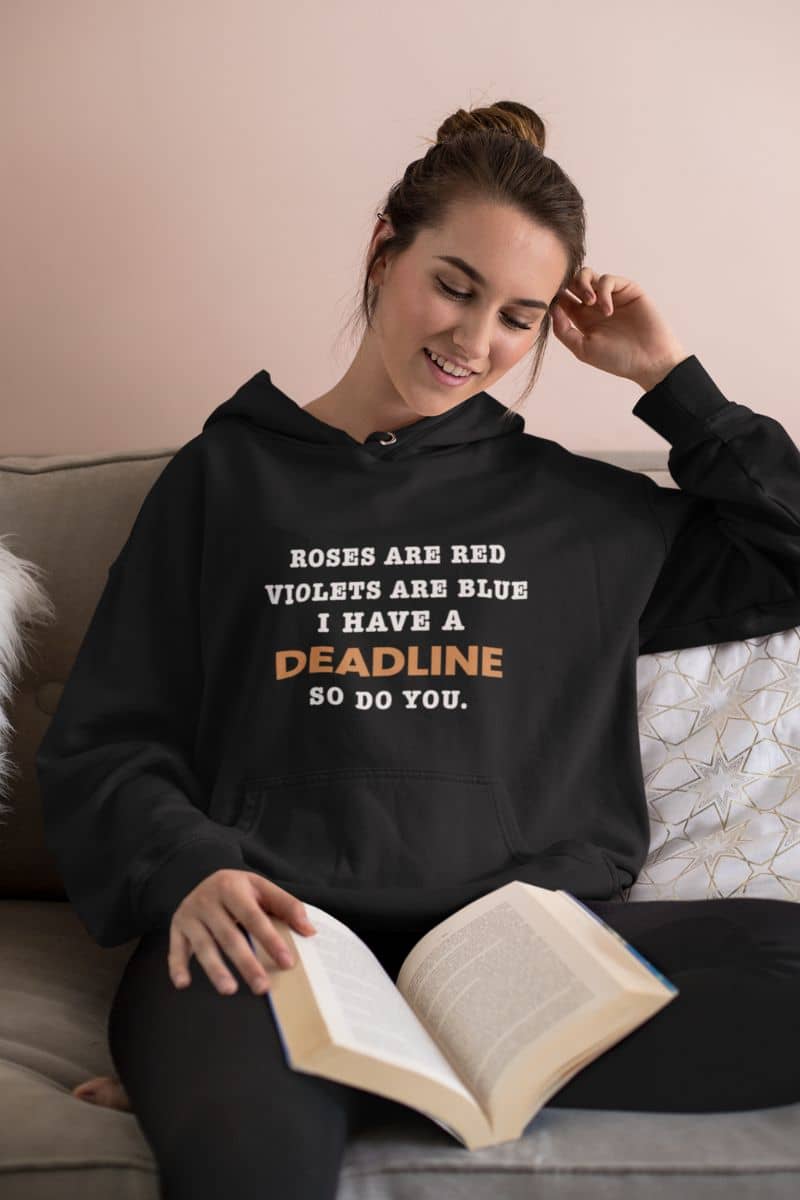 sarcastic work hoodie with a woman wearing a black hoodie with text 'Roes are red, violets are blue. I have a deadline so you do', sitting on a couch with a book.