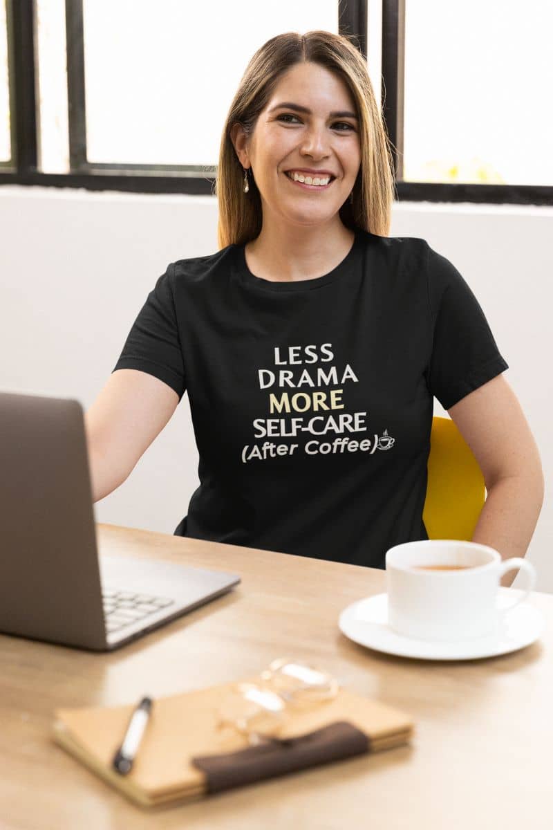 sarcastic work shirt with a woman wearing a black t-shirt with text 'Less Drama, More Self-Care (After Coffee)', sitting at a desk with a laptop and coffee.