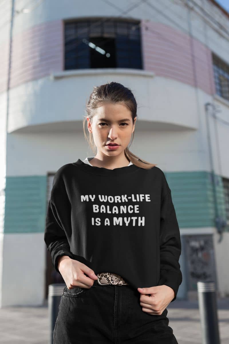sarcastic work sweatshirt with a woman wearing a black sweatshirt with 'My work-life balance is a myth' text in front of a building.