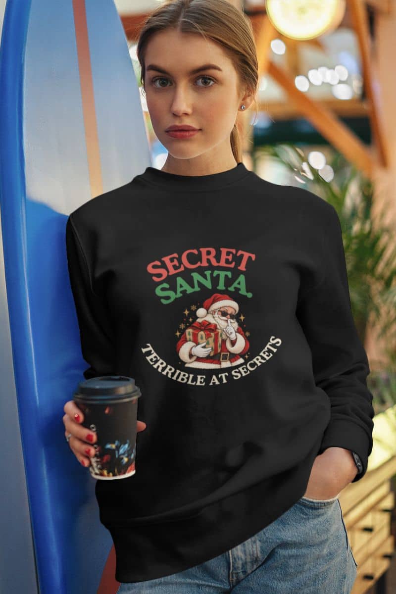 Secret Santa crewneck with a playful Santa in sunglasses holding a Christmas gift, worn by a woman holding coffee.