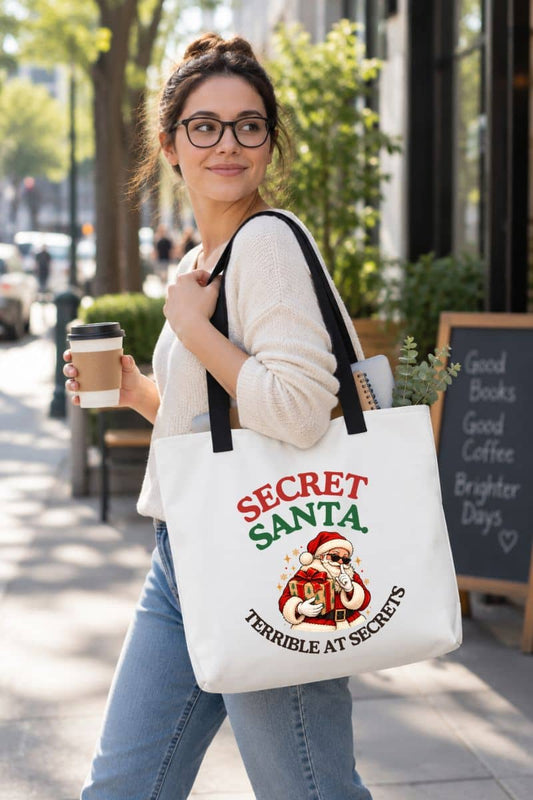 Secret Santa tote bag featuring the “Secret Santa. Terrible at Secrets.” design on a white canvas bag with black handles, carried by a woman holding coffee outdoors.