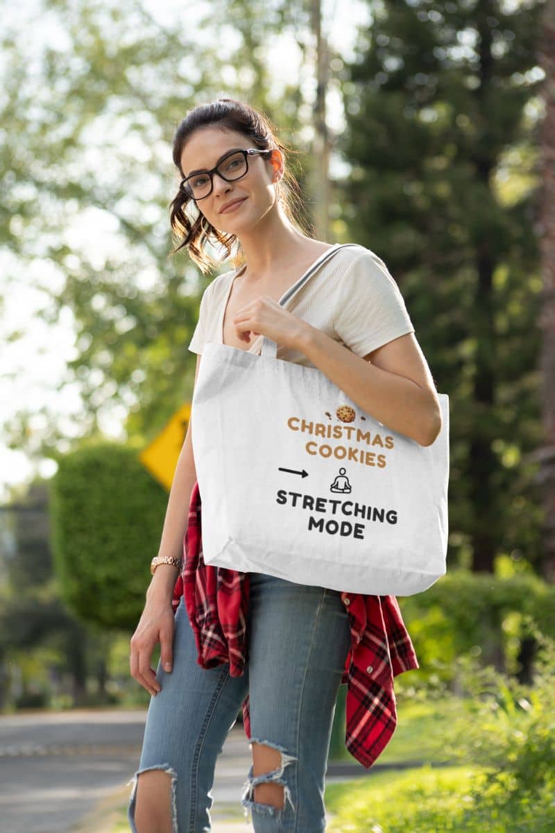 self care canvas tote with a woman holding a tote bag with text 'Christmas Cookies, Stretching Mode' outdoors