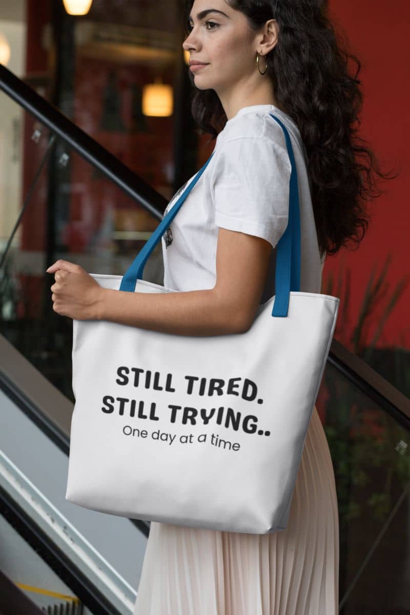 self care canvas tote with a woman carrying a tote bag with motivational text 'Still Tired. Still Trying. One day at a time' in an indoor setting