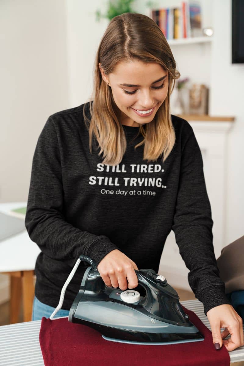 self care quote sweater with a woman ironing clothes wearing a black sweatshirt with a motivational quote 'Still Tired. Still Trying. One day at a time.'