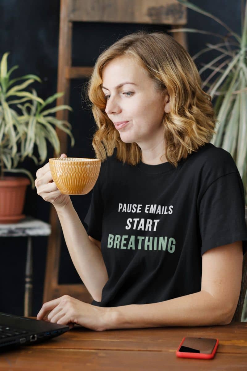 self care shirt with a woman holding a mug with a black t-shirt that says 'Pause Emails, Start Breathing' in a home setting.