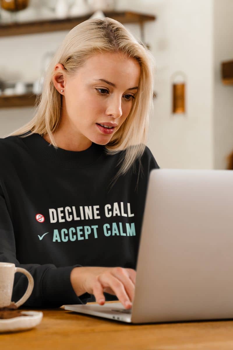 self care sweatshirt with a woman wearing a black sweatshirt with text 'Decline Call, Accept Calm', using a laptop at a table.