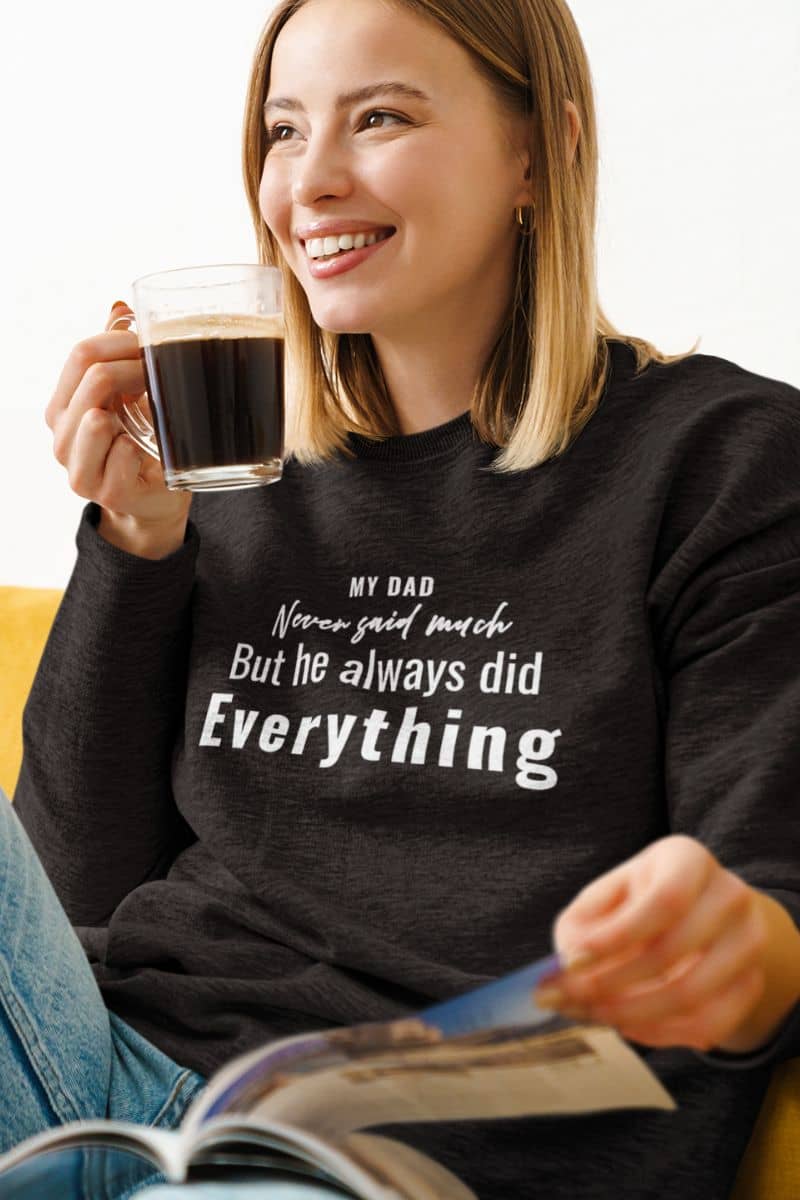 sentimental dad sweatshirt with a woman wearing a black sweatshirt with text 'My Dad Never Said Much, He Always Did Everything, holding a coffee cup and reading a magazine.