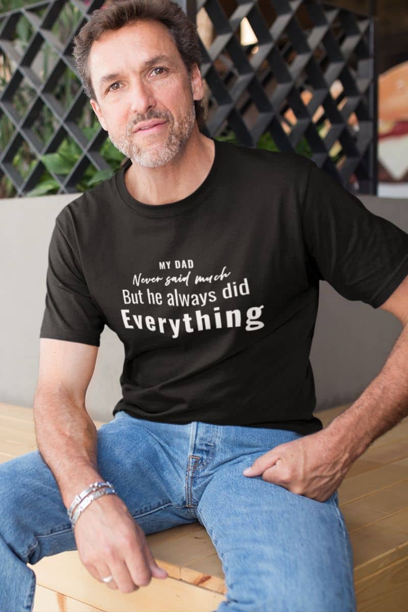 shirt for dad with a man wearing a black t-shirt with text 'My Dad Never Said Much, He Always Did Everything', sitting on a wooden bench.