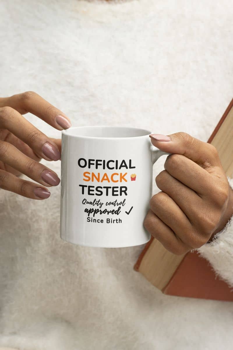 silly coffee mugs with a person holding a mug with 'Official Snack Tester – Quality Control Approved – Since Birth' text against a neutral background