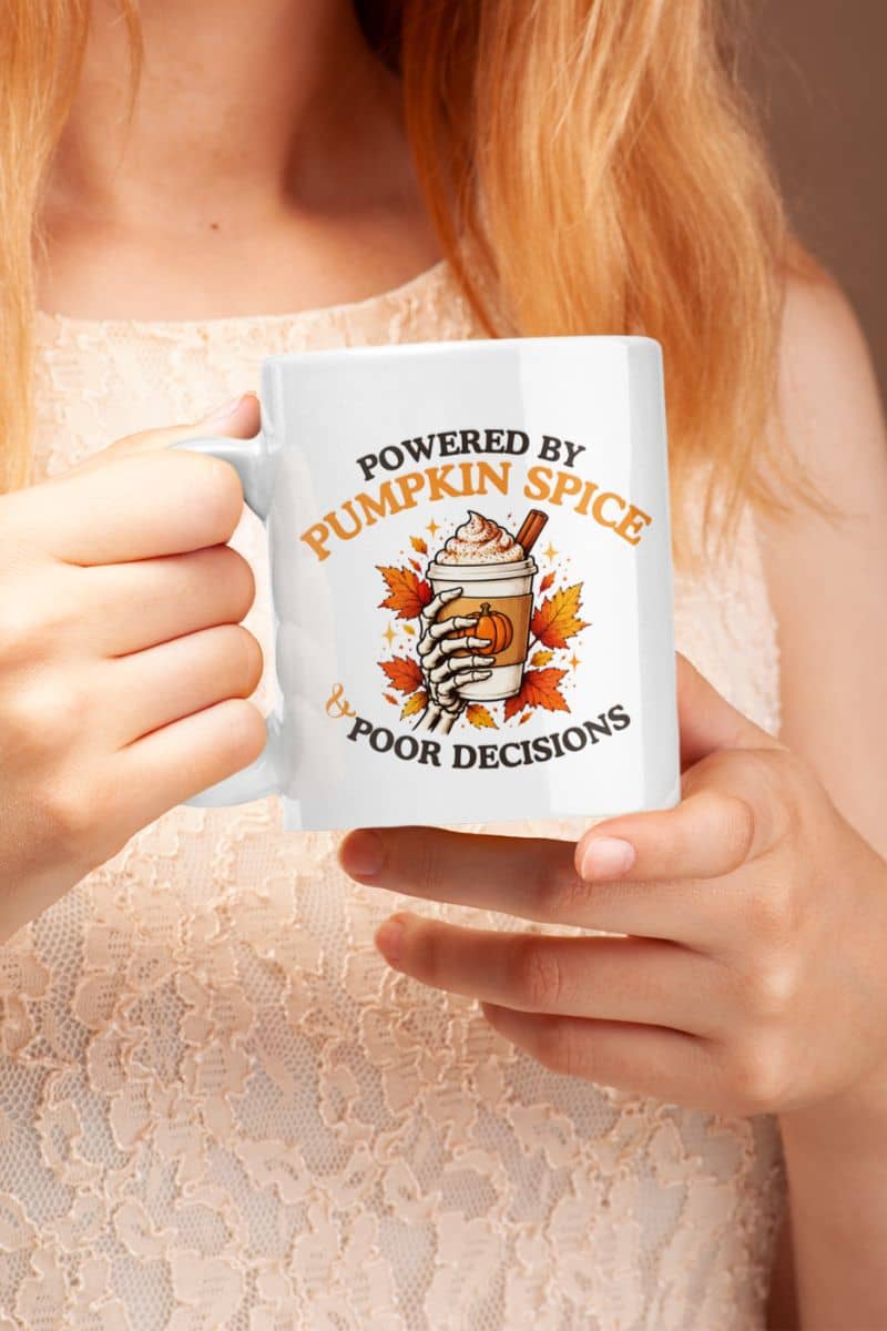 A woman holding a skeleton mug featuring the text 'Powered by Pumpkin Spice & Poor Decisions' and a cup of coffee