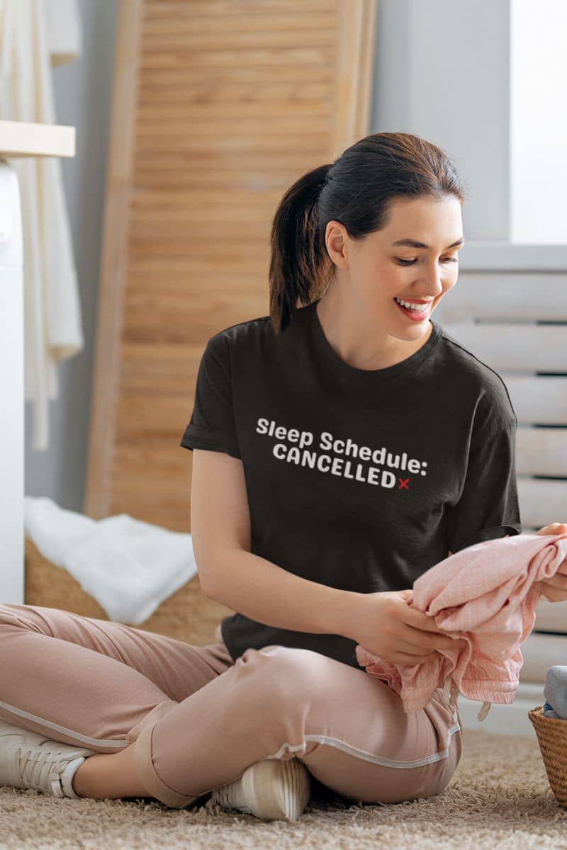 sleep deprived shirt with a woman sitting on the floor holding a pink blanket, wearing a black t-shirt with text 'Sleep Schedule: Cancelled'.