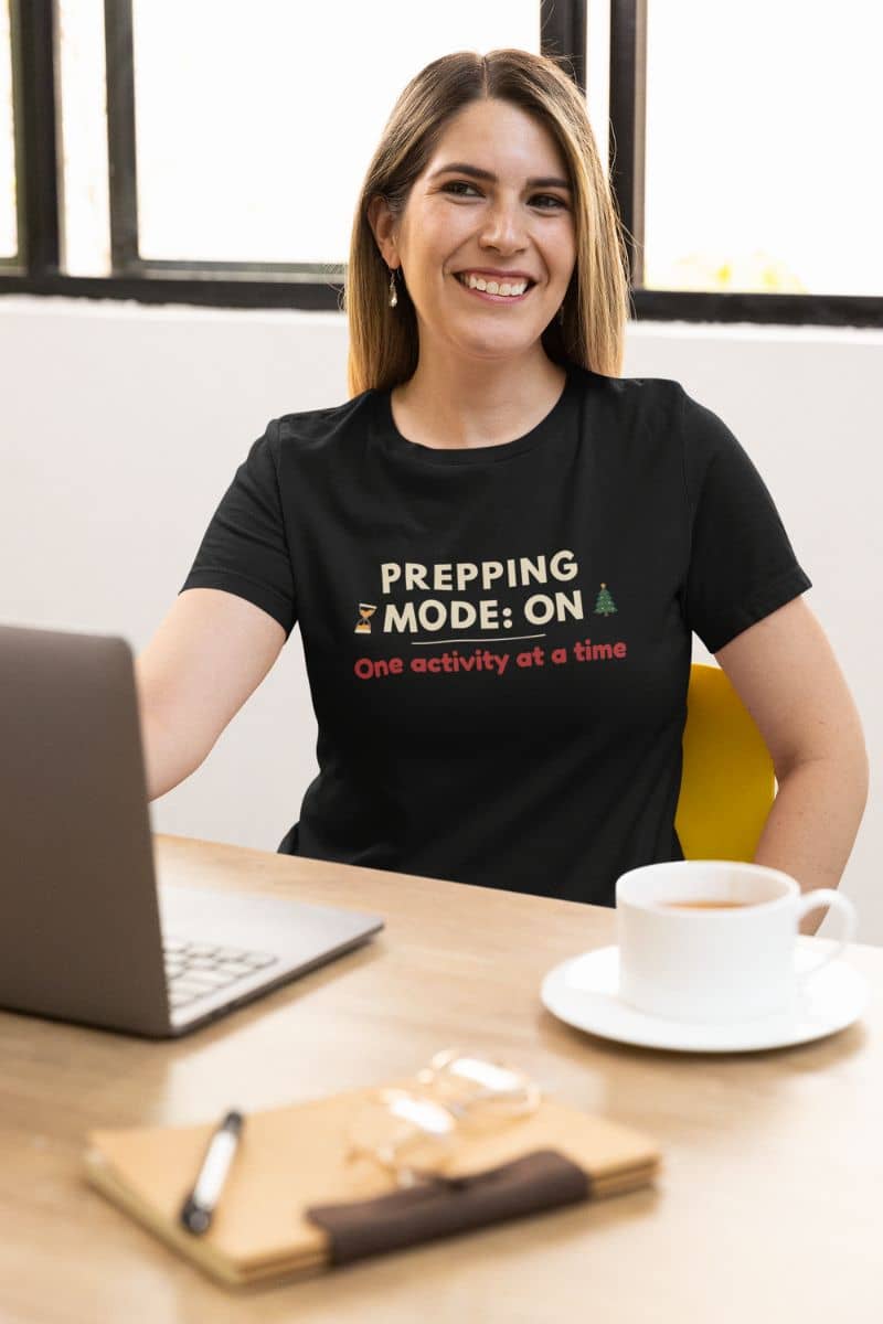 slow living holiday shirt with a woman wearing a black t-shirt with text 'Prepping Mode: ON – One activity at a time', sitting at a desk with a laptop and coffee.