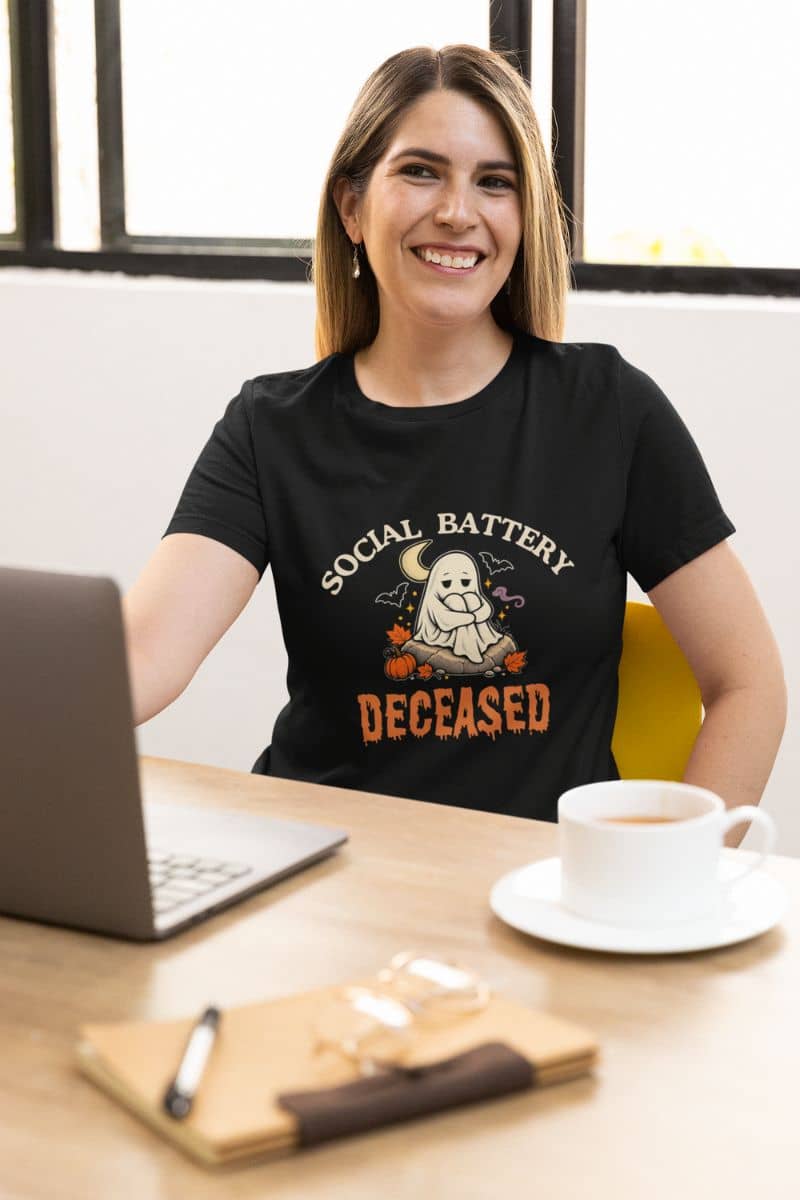 A woman wearing a social battery shirt that reads 'Social Battery: Deceased' with a ghost and pumpkins, smiling at a desk has a laptop, a coffee cup, and a notebook
